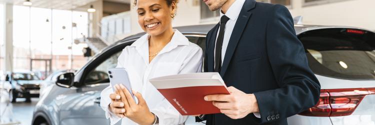 A man and a woman in front of a silver mid-size car in a car dealership while they're both looking at her phone and he's holding papers
