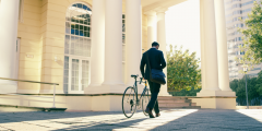 a man turned away from the camera, walking towards a white building. The man is in a suit and holding a bike