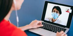 A woman with headphones on talking to a doctor wearing a mask via laptop