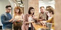 A group of people standing around tablets at an office table while talking and drinking coffee in mugs