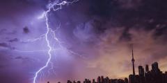 A dark city skyline with clouds overhead and a large bolt of lighting against a purple sky