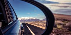 Close-up image of a car's side mirror showing a reflection of the road and mountains and blue sky behind it alongside an image in the foreground