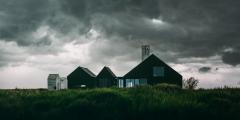 A simple brown house in the countryside atop of tall green grass and beneath a dark and cloudy sky