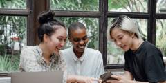 Three young women sitting at a table looking at an electronic device and smiling