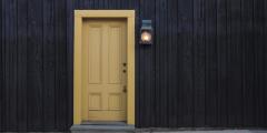 A yellow door against a black wooden wall with a light next to the door