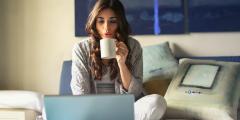 Woman on computer sitting in bed, sipping coffee